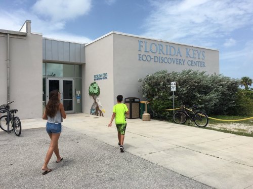 Two children walk towards the Florida Keys Eco-Discovery Center, with bicycles parked nearby and greenery in the background.