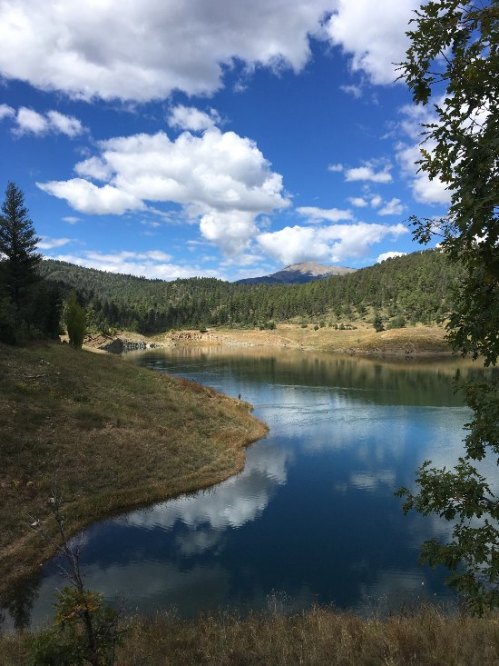 A serene lake surrounded by green hills and trees, reflecting fluffy clouds and a blue sky.