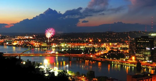 A vibrant city skyline at dusk, with fireworks above and reflections on the water, showcasing lights and bridges.