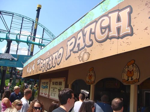 A busy food stand named "Potato Patch" with a roller coaster in the background, filled with people enjoying the day.