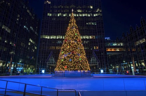 A brightly lit Christmas tree stands in an ice skating rink, surrounded by modern buildings at night.