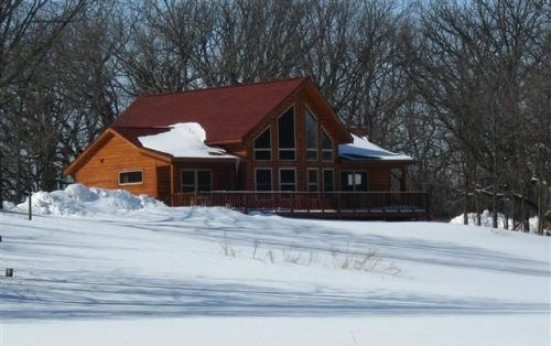 A wooden house with a red roof surrounded by snow and trees on a winter day.