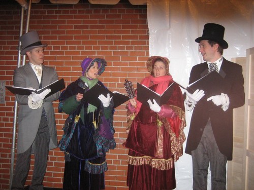 Four performers in Victorian costumes sing together, holding music books, against a brick wall backdrop.
