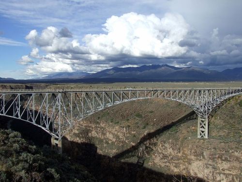 A large metal bridge spans a deep canyon, with mountains and cloudy skies in the background.