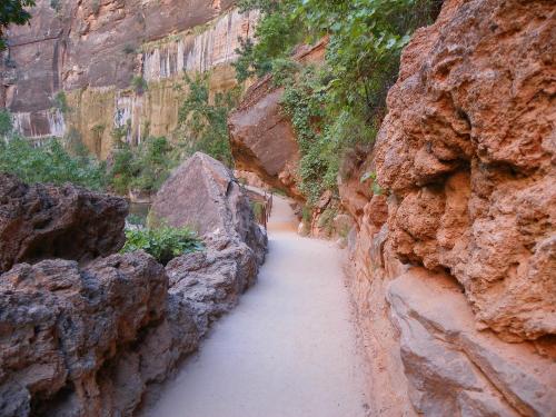 A narrow sandy path winds through rocky terrain, surrounded by lush greenery and towering cliffs.