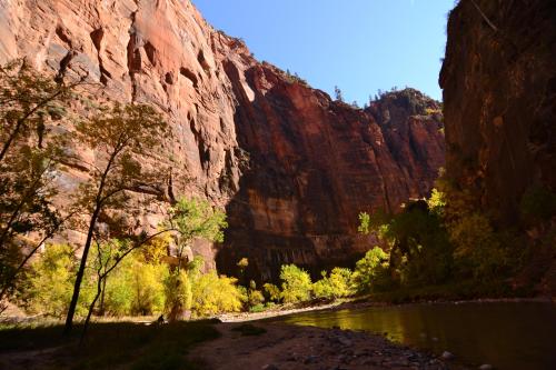 A scenic view of towering red rock cliffs beside a calm river, with vibrant green and yellow trees in the foreground.
