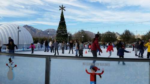 A lively outdoor ice skating rink with a decorated Christmas tree and mountains in the background, filled with skaters.