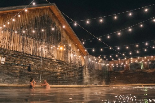 Two people relax in a warm pool at night, surrounded by string lights and a rustic wooden building.