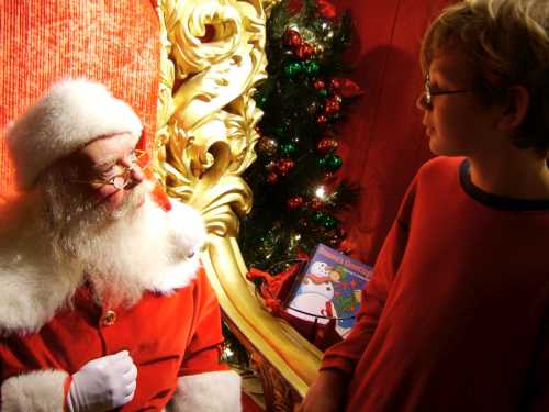 A child in a red shirt talks to Santa Claus in a festive setting with Christmas decorations.