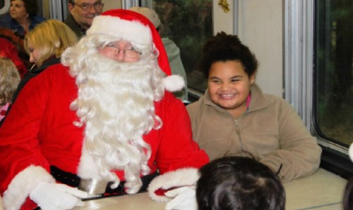 A smiling child sits next to Santa Claus on a train, surrounded by other passengers in a festive setting.