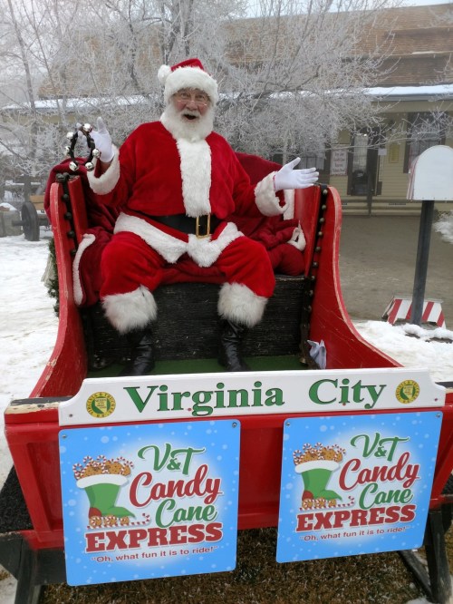 Santa Claus sits in a festive sleigh, smiling and waving, with signs for the Virginia City Candy Cane Express nearby.