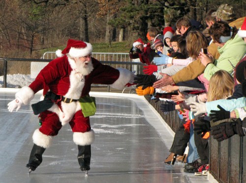 Santa Claus ice skating while children and adults reach out to him, all wearing festive hats and gloves.
