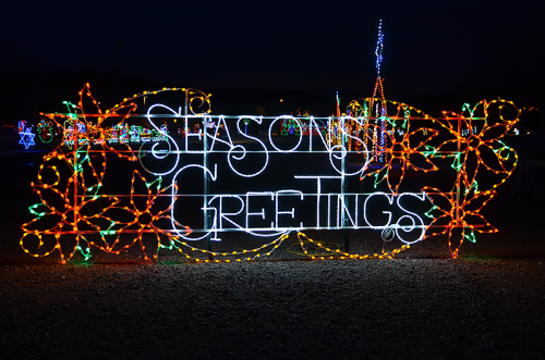 Colorful holiday lights spell out "Season's Greetings" surrounded by festive floral designs against a dark background.