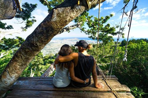 A couple sits on a wooden platform, embracing, with a scenic view of mountains and a city in the background.