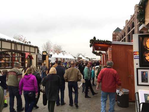 Crowd of people walking through a festive outdoor market with vendor stalls and holiday decorations.