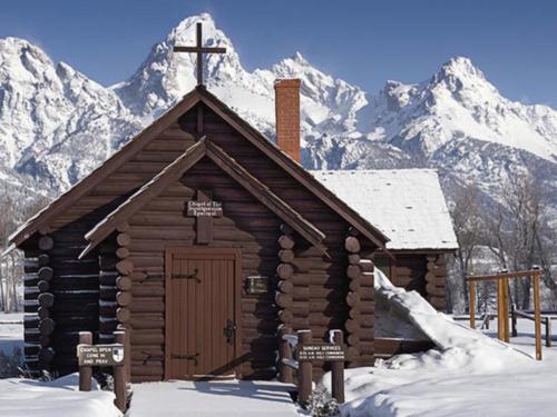 A rustic log chapel with a cross, surrounded by snow and mountains in the background.