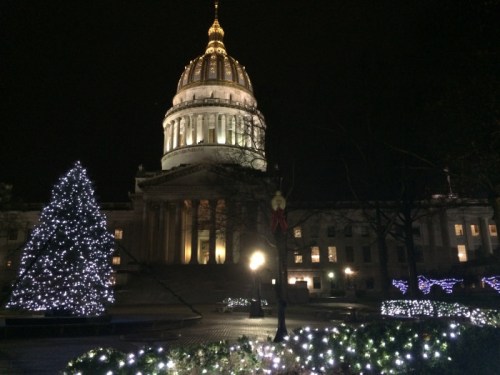 The state capitol building illuminated at night, surrounded by festive lights and a decorated tree.