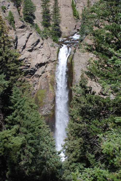 A tall waterfall cascades down rocky cliffs, surrounded by lush green trees and rugged terrain.