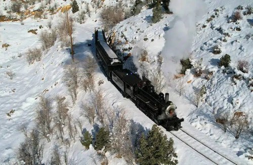 A vintage steam train travels through a snowy landscape, surrounded by trees and mountains. Smoke billows from the engine.