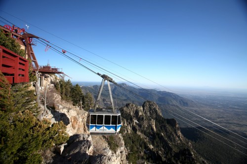 A cable car glides over a mountain landscape, with clear skies and distant valleys visible below.