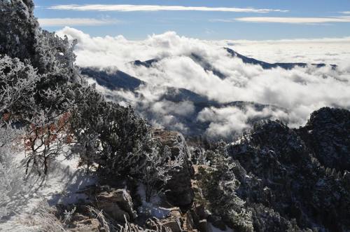 A snowy mountain landscape with clouds rolling over peaks under a clear blue sky.