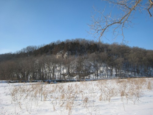 Snow-covered landscape with a hill and bare trees under a clear blue sky.