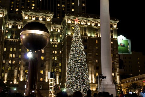 A brightly lit Christmas tree stands in front of a grand building at night, surrounded by festive decorations.
