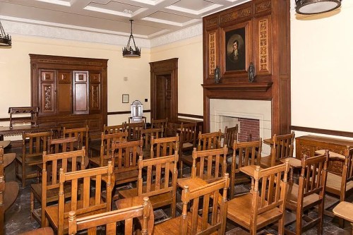 A historic classroom with wooden chairs arranged around a fireplace and a portrait on the wall.