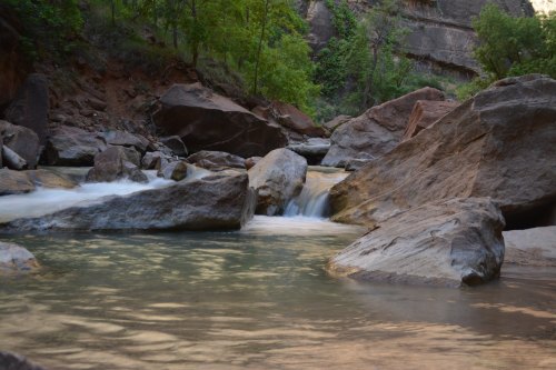 A serene river flows over rocks, surrounded by lush greenery and steep canyon walls.