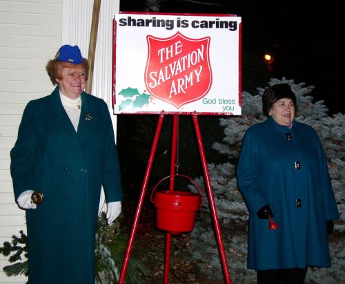 Two women in teal coats stand beside a Salvation Army donation kettle, smiling and holding bells. A sign reads "sharing is caring."
