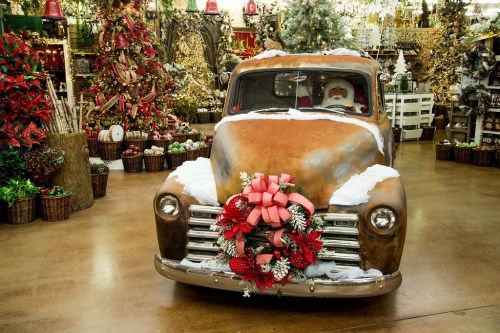A vintage truck decorated with a festive wreath, surrounded by holiday decorations and a Christmas tree.