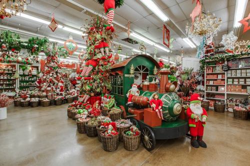 A festive holiday display featuring a train, decorated trees, and Santa figures surrounded by colorful decorations and gifts.
