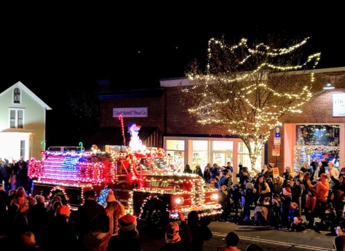 A festive parade scene at night with a decorated fire truck and a crowd enjoying the holiday lights and decorations.