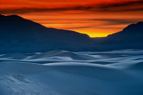 A stunning desert landscape at sunset, with rolling sand dunes and mountains silhouetted against a vibrant orange sky.