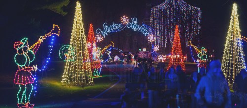 Colorful holiday lights illuminate a festive scene with trees, decorations, and cheerful figures playing instruments.