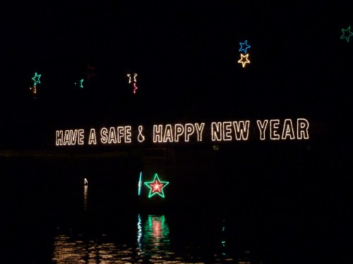Brightly lit sign reads "HAVE A SAFE & HAPPY NEW YEAR" with colorful stars, reflecting on water at night.