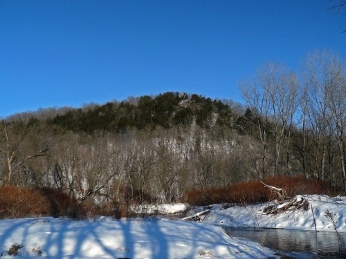 A snowy landscape with a hill in the background, bare trees, and a calm river reflecting the clear blue sky.