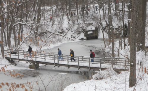 People walking on a wooden bridge over a frozen river, surrounded by snow-covered trees in a winter landscape.