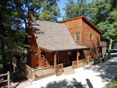 A cozy log cabin surrounded by trees, featuring a porch and a gravel path leading to the entrance.