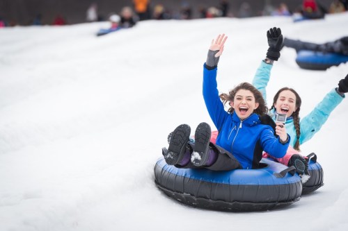 Two girls joyfully tubing down a snowy hill, waving and smiling, with other tubers in the background.