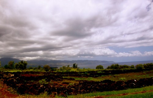A scenic landscape featuring rolling hills, dark volcanic rock, and a cloudy sky over a distant coastline.