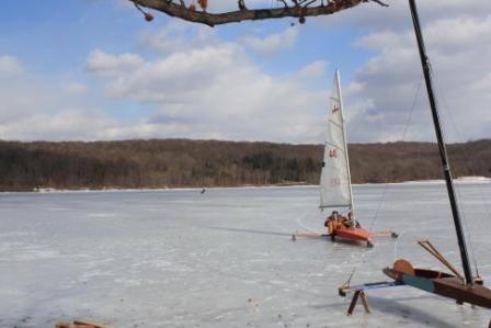 A small sailboat glides across a frozen lake under a cloudy sky, with trees in the background.