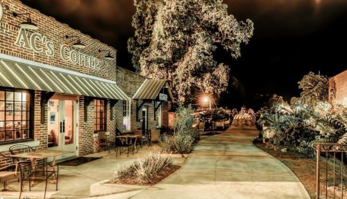 A nighttime view of a coffee shop with outdoor seating, surrounded by trees and a well-lit pathway.