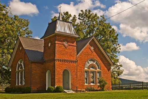 A quaint brick church with a steeple, surrounded by greenery and under a blue sky with fluffy clouds.