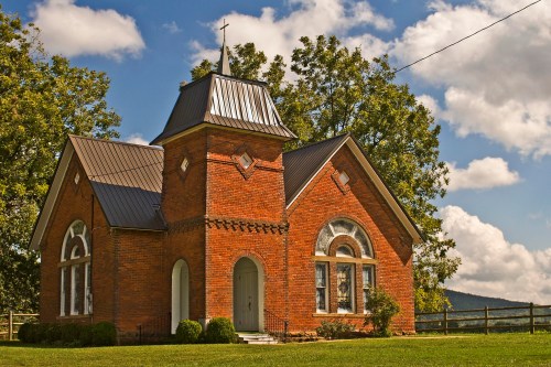 A quaint brick church with a steeple, surrounded by greenery and under a blue sky with fluffy clouds.