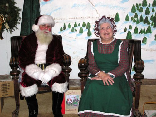 Santa Claus and Mrs. Claus sit together in festive attire, smiling in front of a painted winter scene.