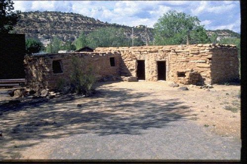 Historic stone building in a desert landscape, surrounded by trees and mountains under a partly cloudy sky.