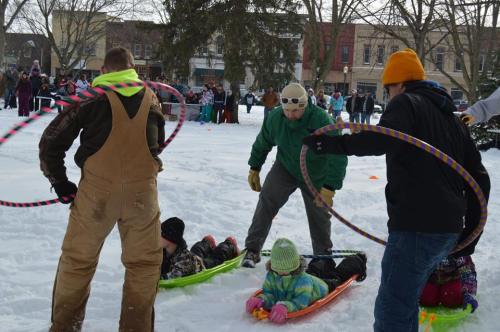 A snowy scene with people sledding and hula hooping, surrounded by a crowd enjoying winter activities.