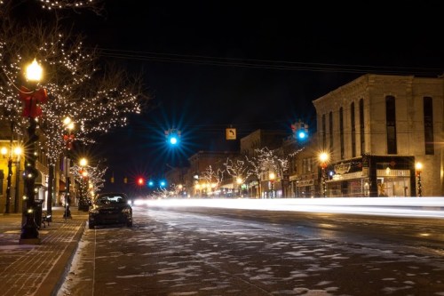 A quiet street at night, lined with festive lights and a blurred car passing by, creating a serene winter atmosphere.