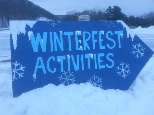 A blue sign with white lettering pointing right, reading "Winterfest Activities," surrounded by snow.